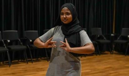 A student performs a dance pose during a drama class on stage, with black curtains and empty chairs in the background